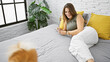 Smiling young hispanic woman lying in bed with her beloved dog, gleefully using her smartphone in the cozy comfort of her bedroom