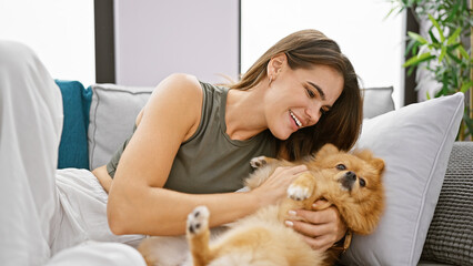Wall Mural - Confident young hispanic woman enjoying a fun laugh, hugging her pet dog while sitting joyfully on her home sofa with a beautiful smile.