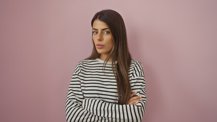 Poster - Confident young hispanic woman with crossed arms posing against a solid pink background, wearing striped shirt