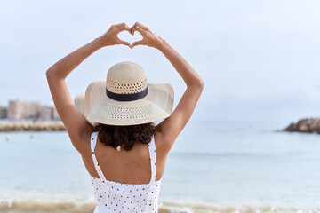 Poster - Young african american woman wearing summer hat doing heart gesture with hands at seaside