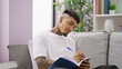© Krakenimages.com - Handsome young latin man sitting on his living room sofa, engrossed in writing on a notebook, ideas flowing, doubts easing, studying while tattoo peeking, home interior setting background.