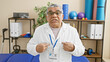 © Krakenimages.com - A mature man in a clinic wearing a lab coat and id badge stands confidently in a rehabilitation room with exercise equipment.