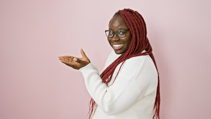 Poster - Portrait of a smiling african american woman with braids, wearing glasses and presenting something against a pink background.