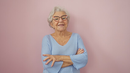 A smiling senior woman with grey hair and glasses stands arms crossed in front of a pink wall wearing a blue sweater.