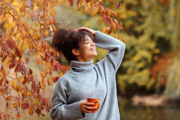 Wall Mural - Young happy smiling mixed-race woman with coffee cup in autumn nature, pleased african american female with curly hair in knitted sweater