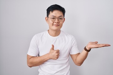 Sticker - Young asian man standing over white background showing palm hand and doing ok gesture with thumbs up, smiling happy and cheerful