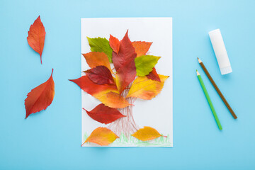 Wall Mural - Tree shape created from colorful leaves on white paper, glue stick and pencils on light blue table background. Pastel color. Making autumn decorations. Closeup. Top view.