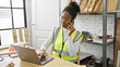 A focused african american woman multitasking in a carpentry studio, on the phone while using a laptop.
