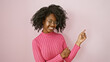 Smiling african woman in pink sweater points happily against a plain indoor background, exuding beauty and confidence.