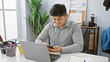© Krakenimages.com - A young asian man smiles while using a smartphone at his modern office desk, portraying a professional, work environment.
