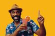 happy bearded mid adult african american man wearing Hawaiian shirt and hat smiling and orange juice cocktail pointing up and looking away at copy space on yellow background