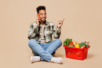 Wall Mural - Full body young man wear grey shirt hold red basket bag with food products talk speak mobile cell phone point finger aside isolated on plain beige background. Delivery service from shop or restaurant.