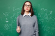 Young strict serious smart teacher woman wears grey casual shirt glasses hold pointer look camera isolated on green wall chalk blackboard background studio. Education in high school college concept.