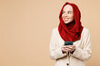 Young Arabian Asian Muslim woman wear red abaya hijab suit clothes hold use mobile cell phone look aside on area isolated on plain beige background studio. UAE middle eastern Islam religious concept.