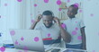 Image of pink spots floating over african american father and son using phone headset and laptop