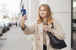 Laughing young blonde woman taking selfie on city background. 30s businesswoman with coffee cup smiling and using smart phone. Urban lifestyle concept. High quality photo