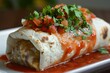 Close-up of a delicious burrito smothered in tomato sauce and garnished with fresh herbs on a white plate.