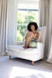 Biracial young woman sitting in white armchair, looking thoughtful at home