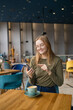 Portrait of happy beautiful 30s blonde woman holding mobile and looking at smartphone while drinking coffee in cafe indoors. Businesswoman drinking coffee and smiling. Girls using an application in