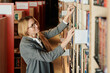 © Clique Images - Happy young woman with long blond straight hair standing in front of large stack of shelves in library and looking for book
