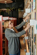 © TrueFrame Collective - Side view of young blond woman in smart casualwear and eyeglasses seeking for book on shelf in modern library or shop