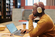 © TrueFrame Collective - Side view of pensive guy in headphones sitting by table in front of laptop with blank screen in modern library