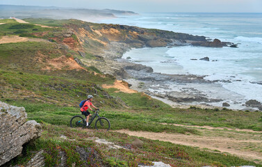 Wall Mural - nice senior woman riding her electric mountain bike at the rocky and sandy coastline of the atlantic ocean in Porto Covo, Portugal, Europe