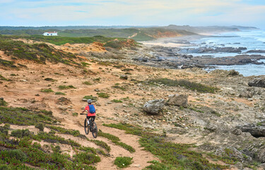 Wall Mural - nice senior woman riding her electric mountain bike at the rocky and sandy coastline of the atlantic ocean in Porto Covo, Portugal, Europe