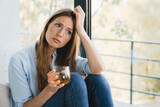 Fototapeta  - Sad stressed young woman thinking about bad memories drinking juice by the window alone. Confused depressed female girl lady drinking wine and having nostalgic feelings