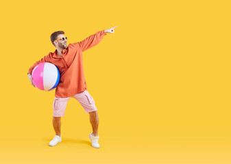 Wall Mural - Happy man with inflatable sea ball pointing finger to copy space. Full length portrait of happy young man wearing summer casual clothes and sunglasses having fun on isolated yellow studio background