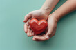Hands cradle a glossy red heart isolated on pastel green background, symbolizing care, love, and protection in a gentle gesture