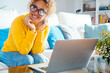 Attractive woman sitting on white sofa at home smiles, looks to her right in front of her laptop. Happy female with yellow sweater spends good time relaxing at home. Leisure and relaxation concept
