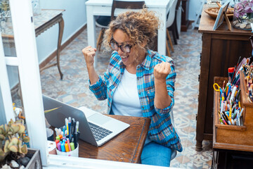 Wall Mural - Attractive woman sitting on chair facing her laptop rejoices, happy about her sale. Happy female does not contain her joy for the money received. Home business and business concept