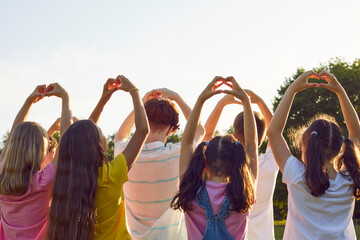 Back view of a group children making heart shape gesture standing in the summer park at sunset. Thankful kids friends doing love sign raising their hands up in nature. Child protection concept