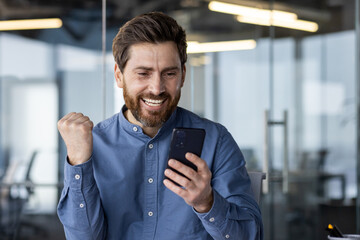 Wall Mural - Happy and smiling young man sitting in office modern center and looking at phone, enjoying success and showing victory gesture with hand. Close-up photo