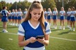 high school cheerleader texting with smart phone on sunny football field