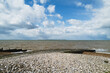Oyster shells on Whitstable beach next to a slipway with a wooden groyne to the left. The clouds are dramatic. The isle of Sheppey can be seen on the horizon.