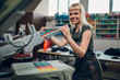 Smiling printing shop worker screen printing t -shirt on machine.