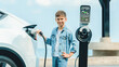 Little boy recharging eco-friendly electric car from EV charging station. EV car road trip travel by the seashore by alternative vehicle powered by clean renewable and sustainable energy. Perpetual