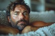 Close-up of a bearded man with deep blue eyes lying wrapped in a textured grey blanket, looking contemplative