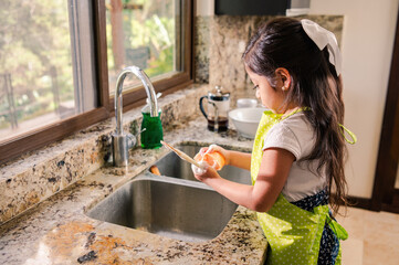 little latina girl with a green apron helps at home washing dishes.