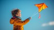 Young boy in bright orange jacket flying a vibrant kite on a clear blue sky day, concept of childhood joy and outdoor play