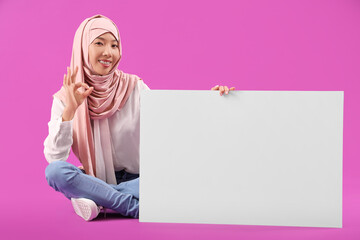 Canvas Print - Beautiful young Asian woman in hijab with blank poster sitting on purple background