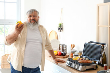 Mature man with bell pepper cooking tasty sausages and vegetables on modern electric grill at table in kitchen