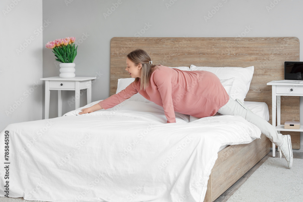 Pretty young woman making bed in light bedroom