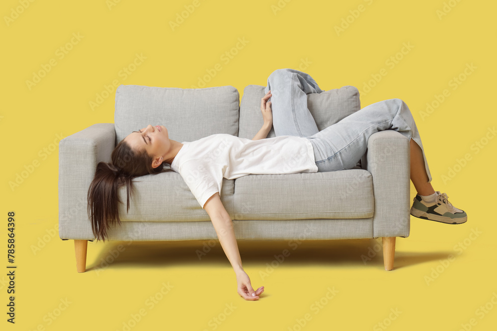 Tired young woman lying on grey sofa against yellow background