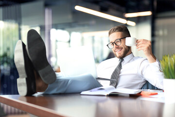 Handsome businessman sitting with legs on table and drinking coffee in office