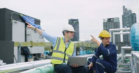 Canvas Print - engineers manager and worker sitting on rooftop review plans on a laptop pointing to something at construction site with high-rise buildings