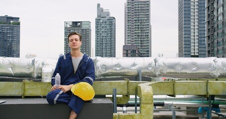 Wall Mural - A engineer worker man in uniform sits on a bench and smile looking to camera at rooftop construction site. The scene takes place in a city with tall buildings in the background