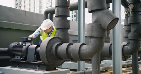 Canvas Print - A Engineer man looking inspecting maintenance insulated pipelines valve pump control on the roof at an industrial site. He is wearing a hard hat and safety vest